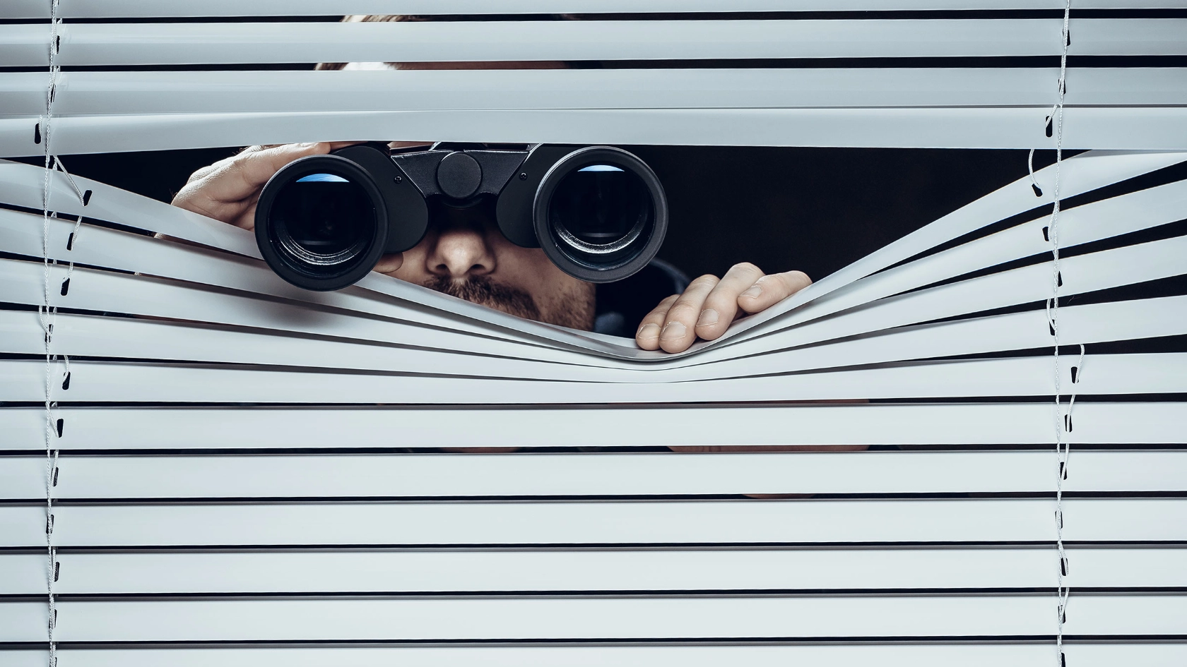 Ein Mann späht offenbar heimlich mit einem Fernglas durch eine Jalousie