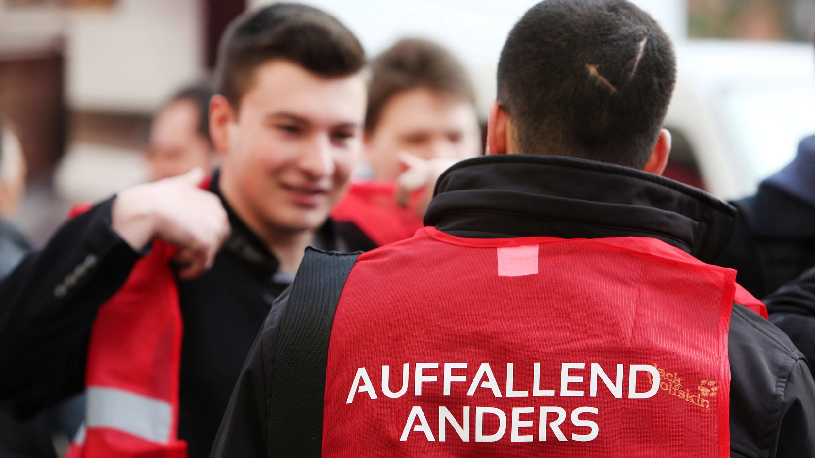 Junger Mann mit roter Weste mit der Aufschrift "Auffallend anders" von hinten.