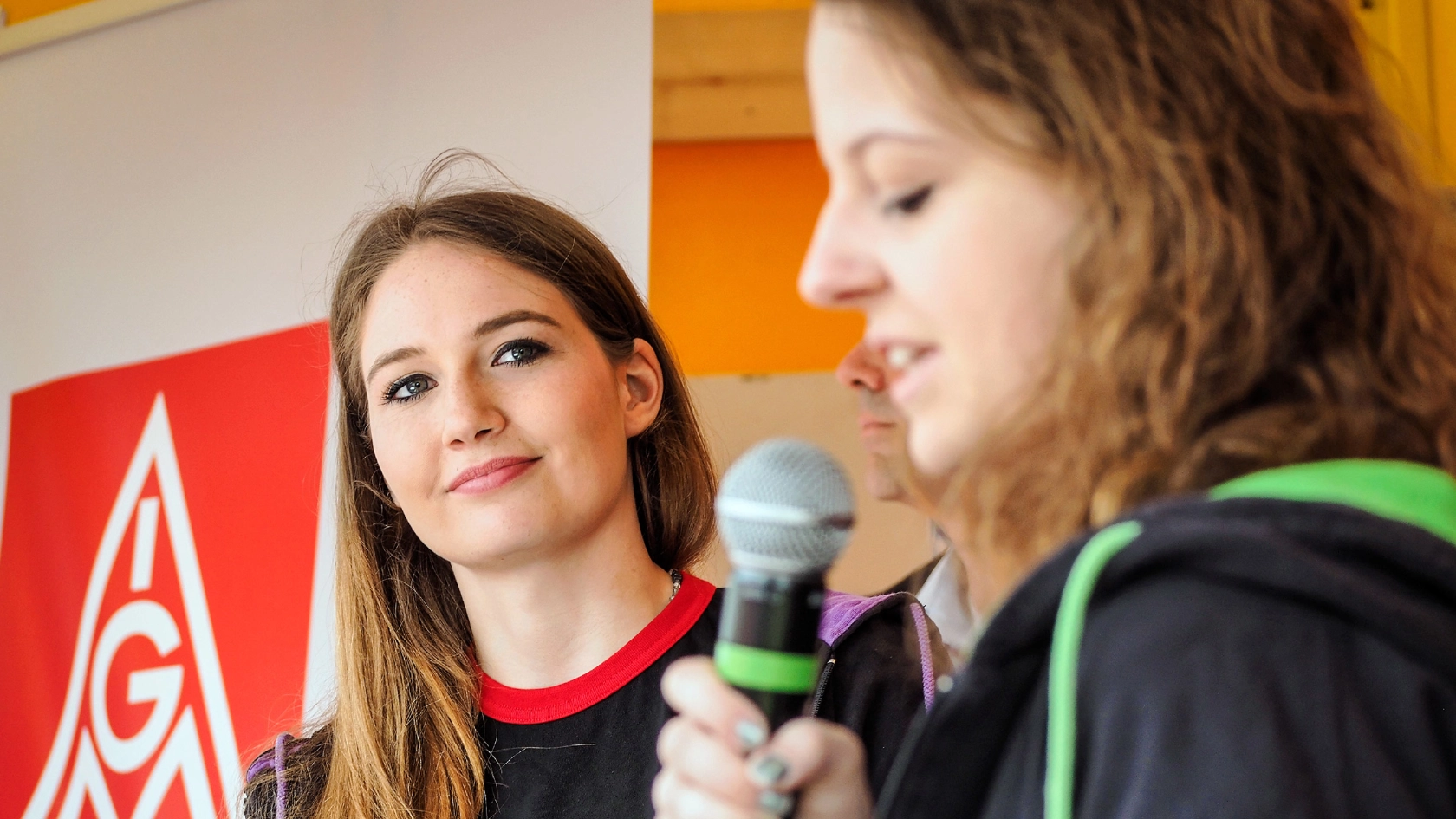 Zwei junge Frauen stehen auf einer Bühne und eine spricht ins Mikro - im Hintergrund eine IG Metall-Fahne.