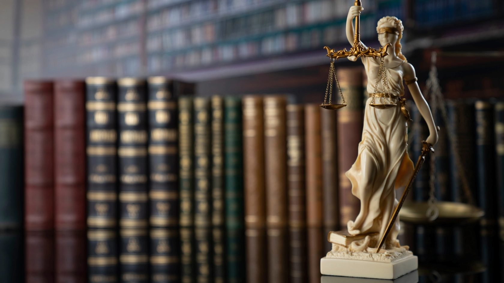 Eine Miniaturstatue von Justizia, welche eine Waage hält steht vor einer Reihe von Rechtsbüchern im Hintergrund