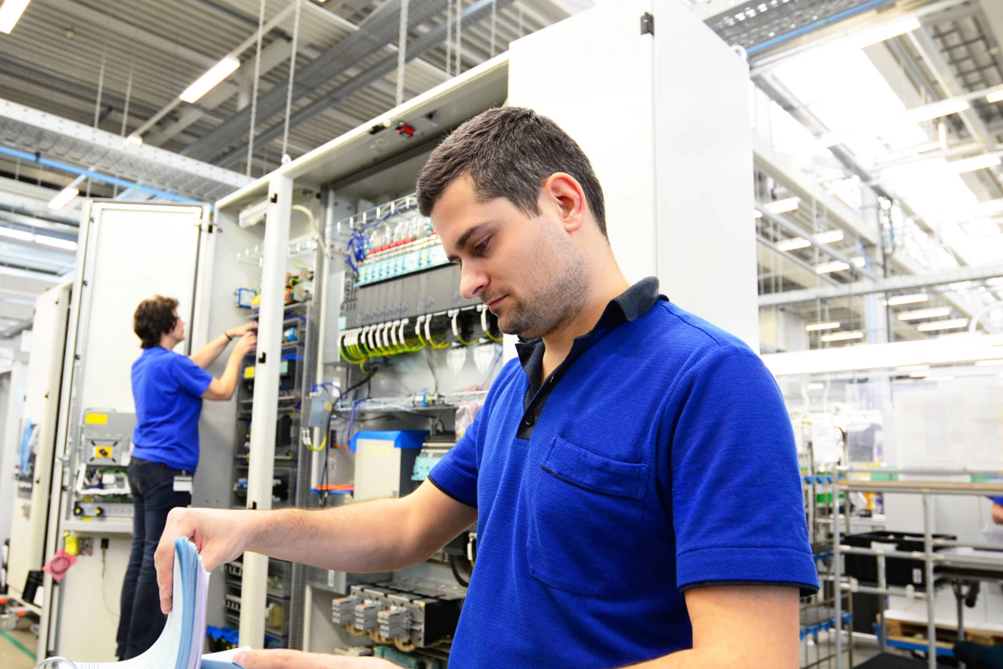 Ein Mann schaut in einen Aktenordner. Im Hintergrund arbeitet eine Frau an Elektronik.