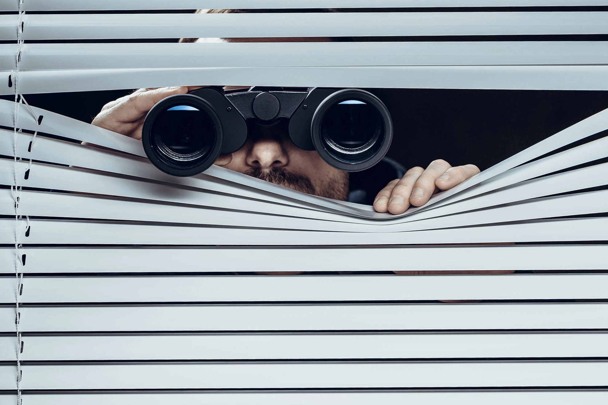 Ein Mann späht offenbar heimlich mit einem Fernglas durch eine Jalousie