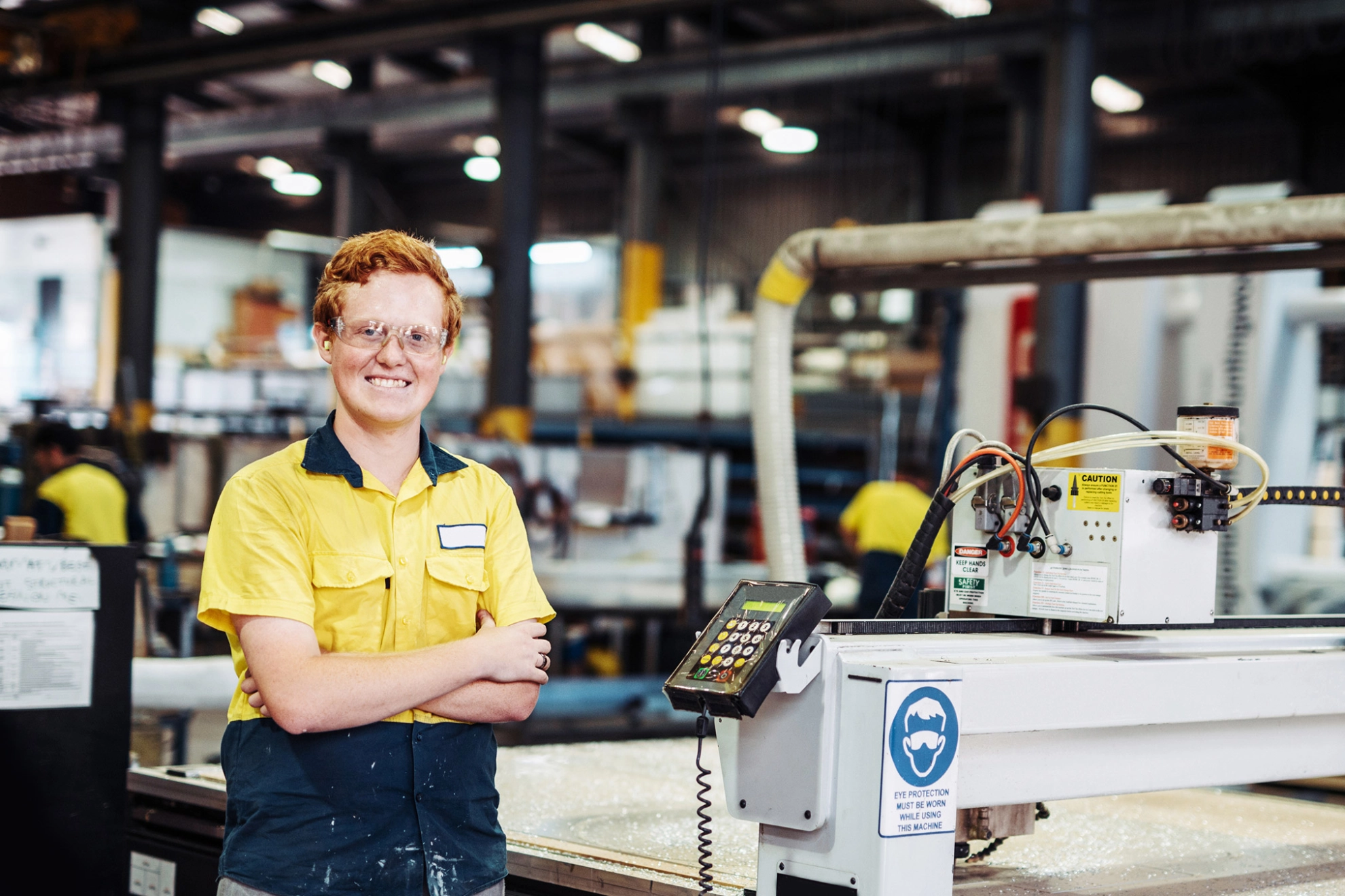 Ein junger Mann mit gelbem Hemd und roten Haaren steht lächelnd neben einer Maschine in einer Fabrik.