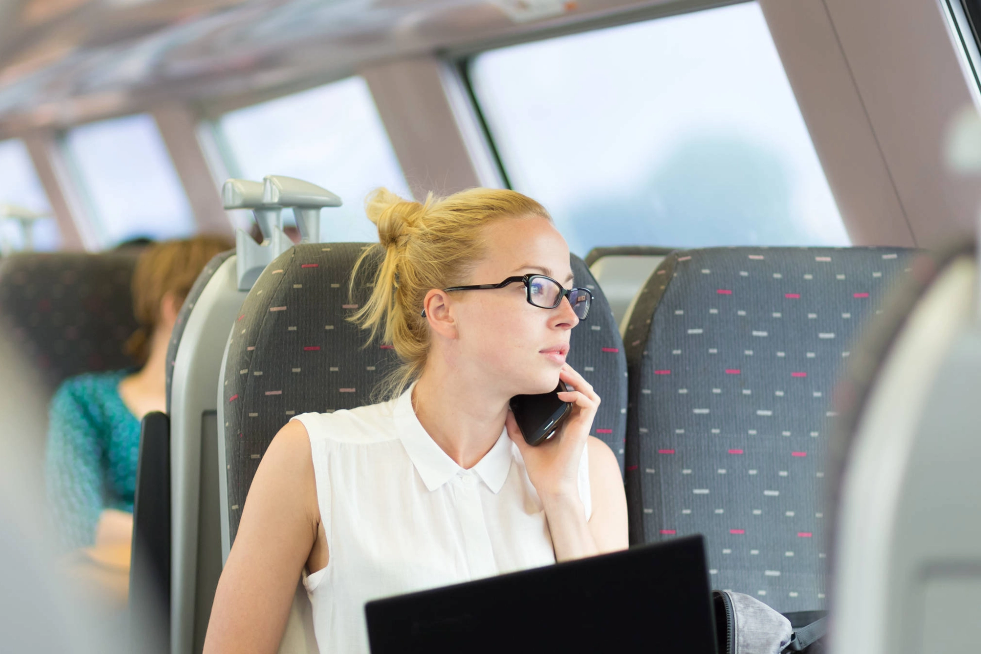Eine junge Frau sitzt mit einem Notebook auf den Knien im Zug und telefoniert.