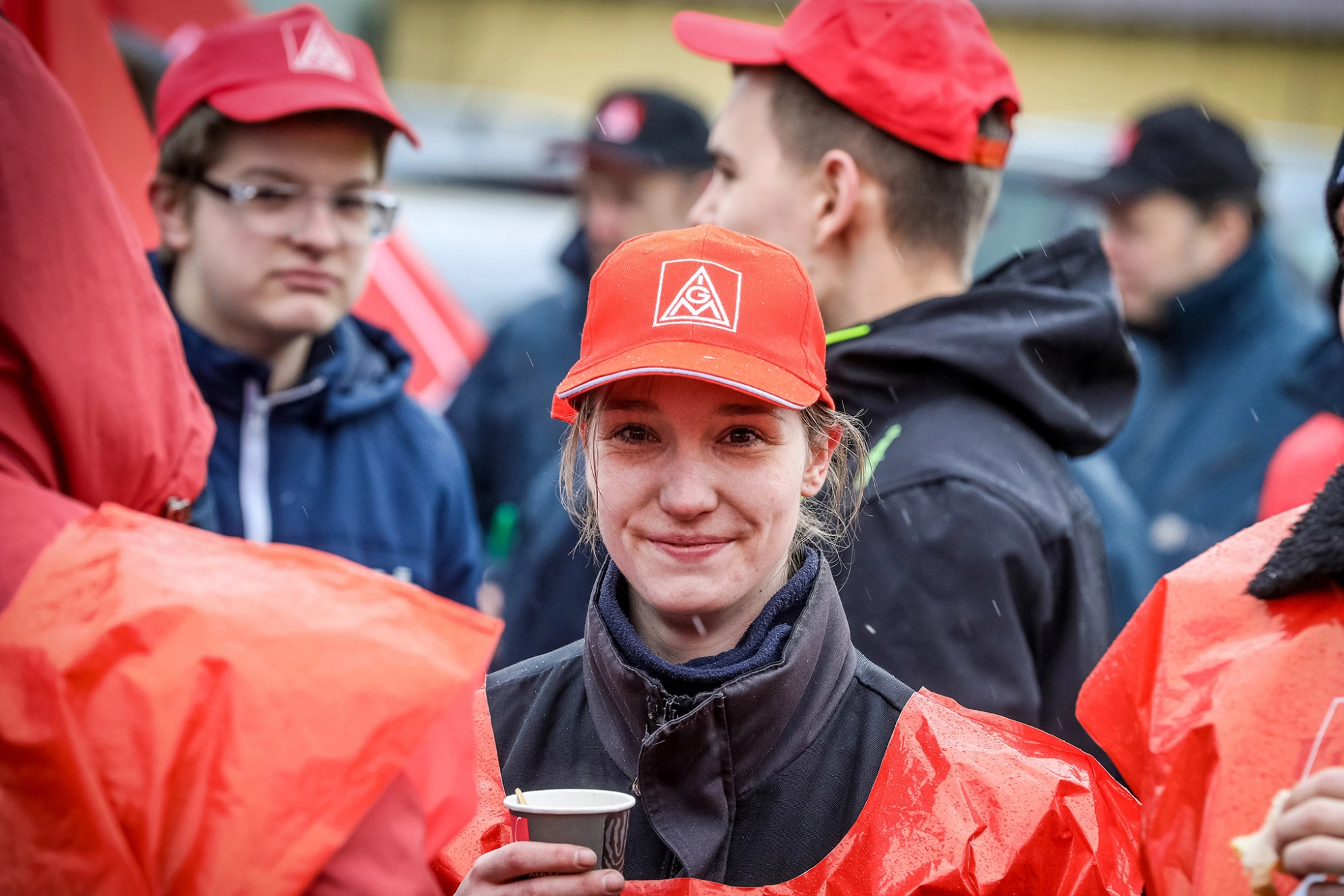 Junge Frau mit einem Kaffeebecher, Warnweste und IG Metall-Kappe.