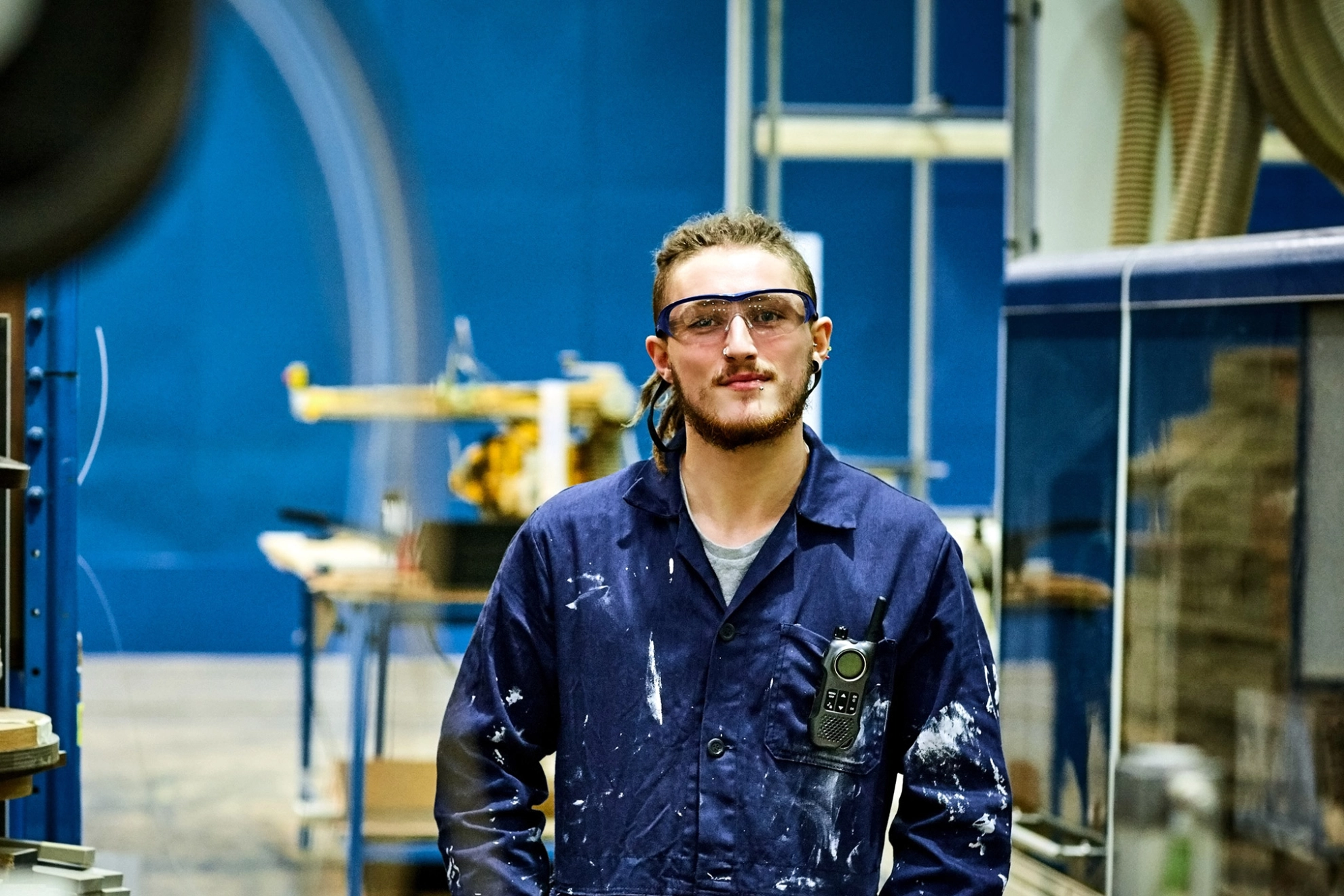 Ein junger Mann im Blaumann mit Schutzbrille und Dreadlocks steht in einer Werkhalle und blickt lächelnd in die Kamera.