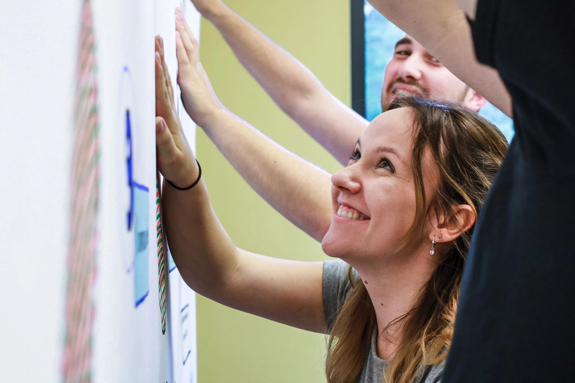 Junge Frau steht während eines Seminars mit weiteren Personen an einer Metaplanwand und schaut sich lächelnd die Aufschrift an.