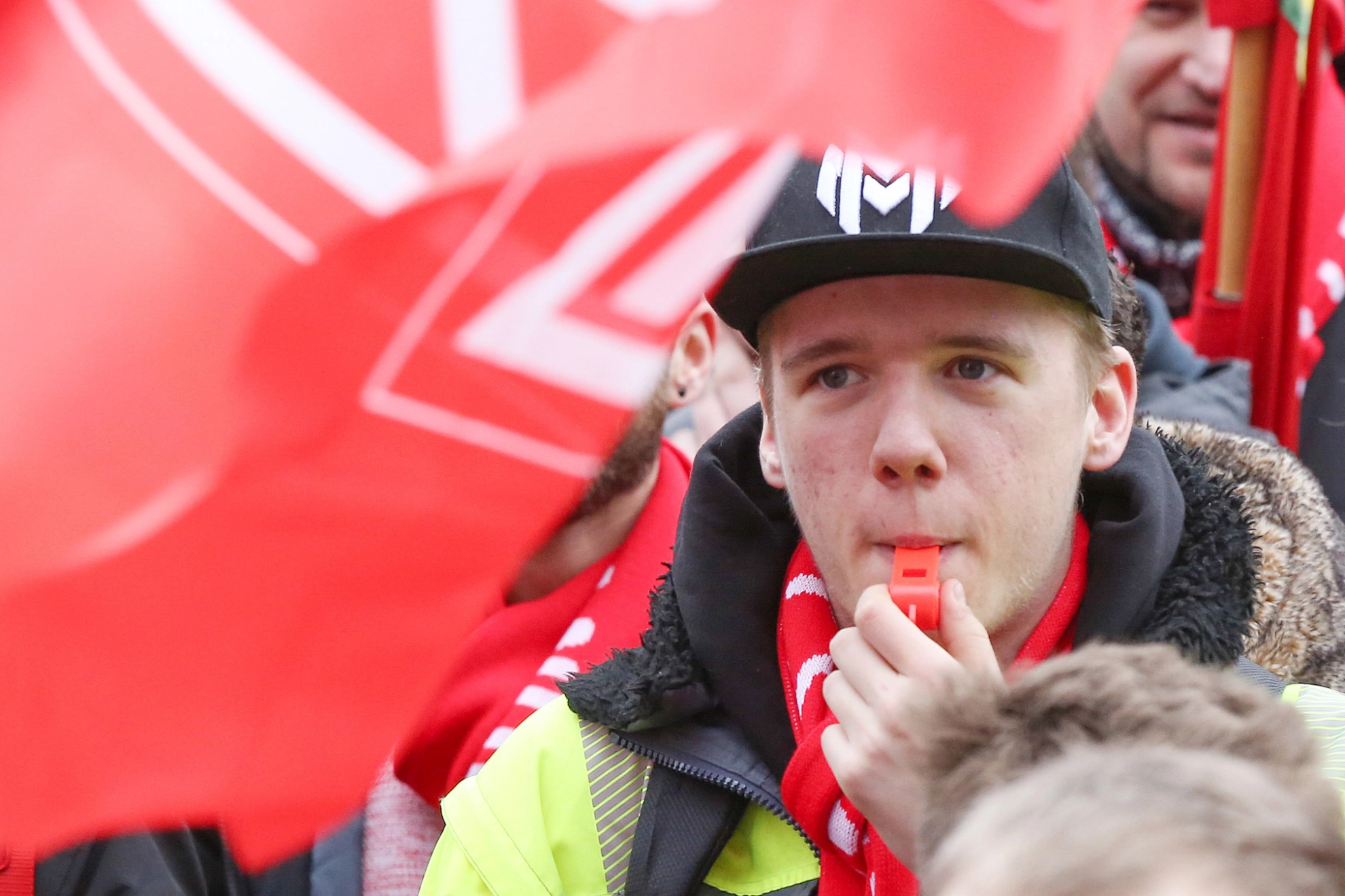 Junger Mann zwischen IG Metall-Fahnen pustet bei einem Warnstreik während der Tarifrunde in eine Trillerpfeife.