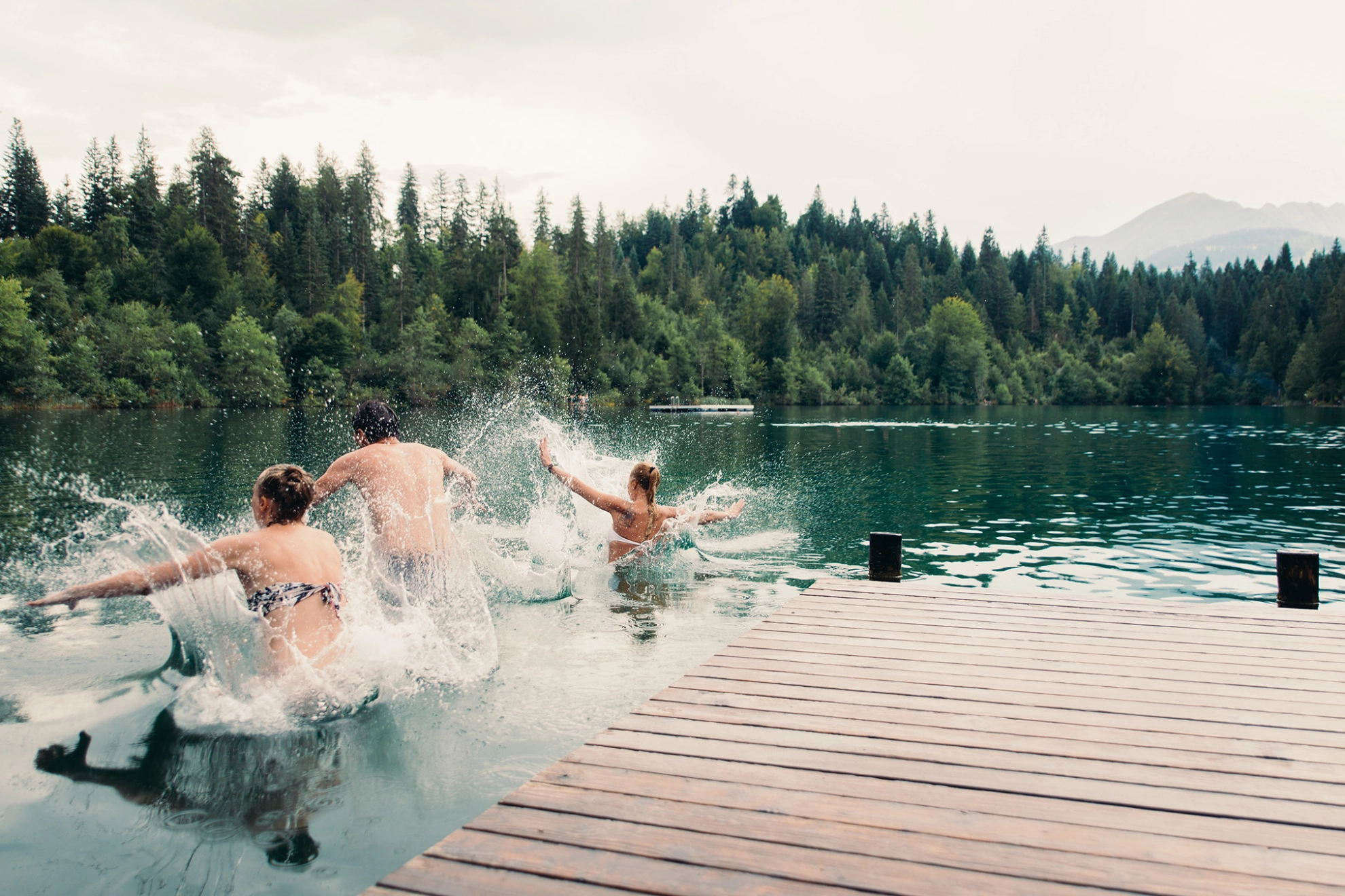 Drei Menschen springen von einem Holzsteg in das Wasser eines Sees