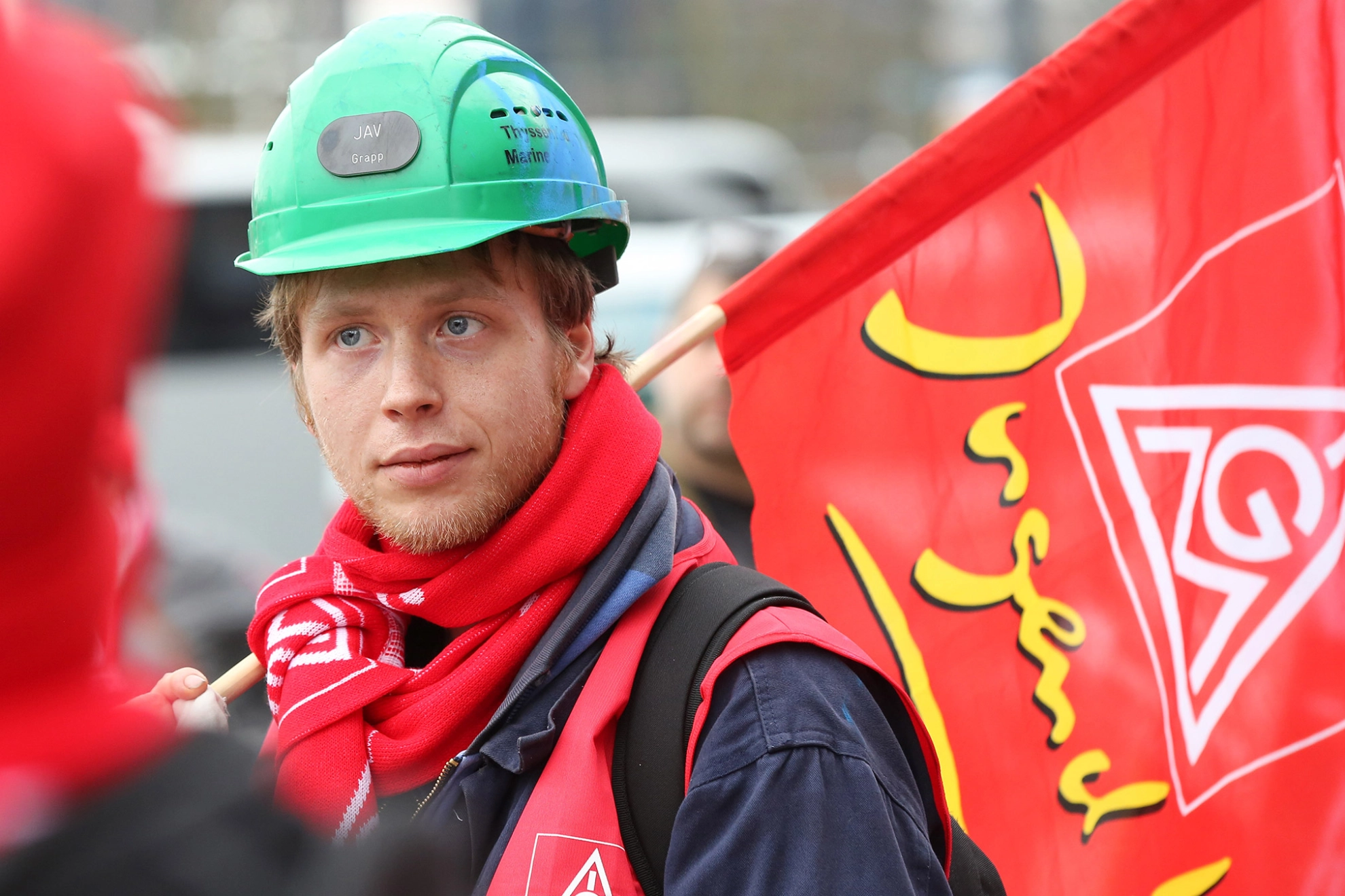 Junger Aktiver mit IG Metall-Fahne und IG Metall-Schal auf einer Demo.