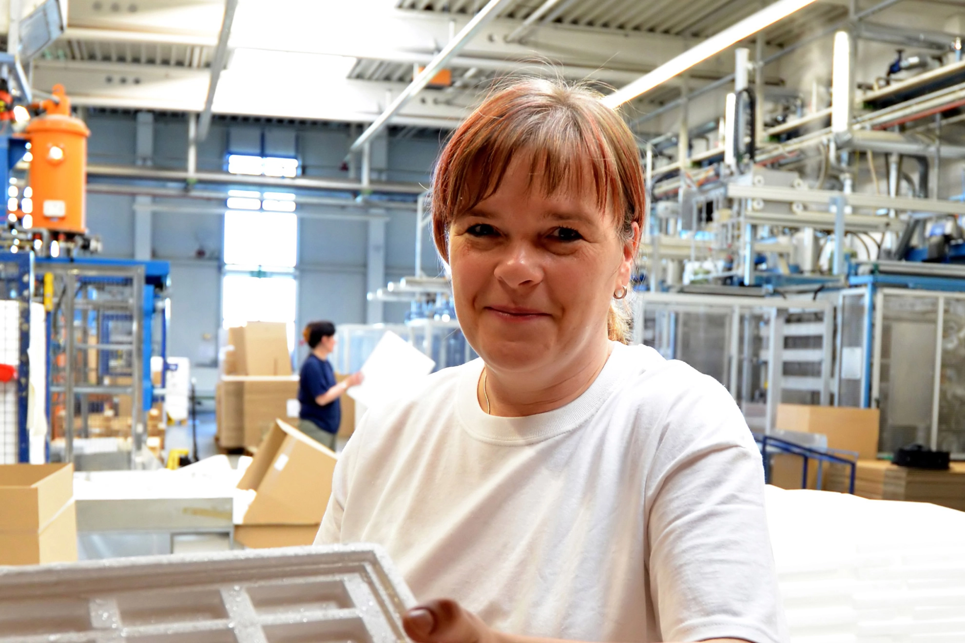 Eine Frau in einer Werkhalle hält eine Styroporkomponente in der Hand und lächelt in die Kamera.