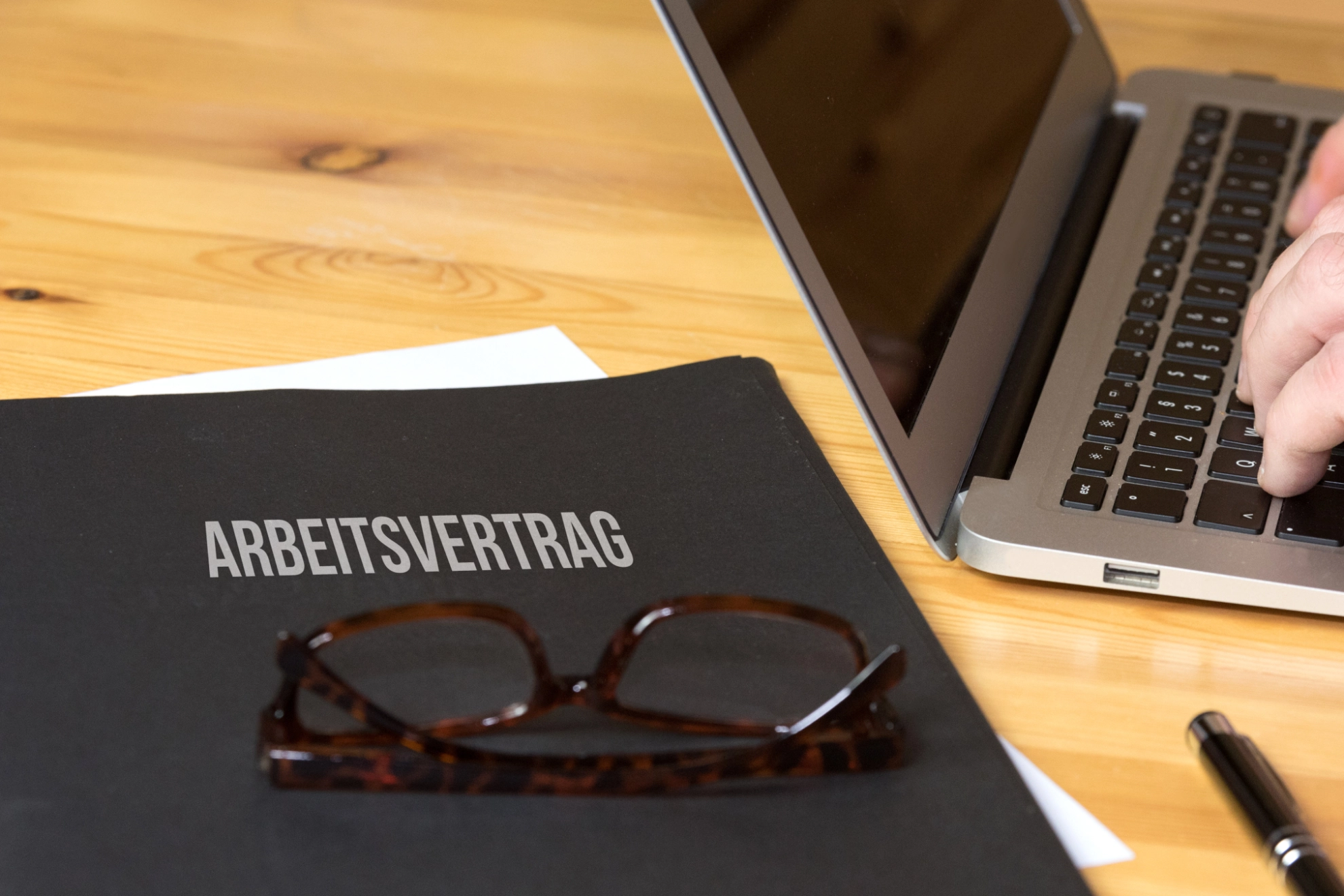 Ein Arbeitsvertrag liegt auf einem Schreibtisch, darauf liegt eine Brille, daneben steht ein Laptop, an dem jemand arbeitet