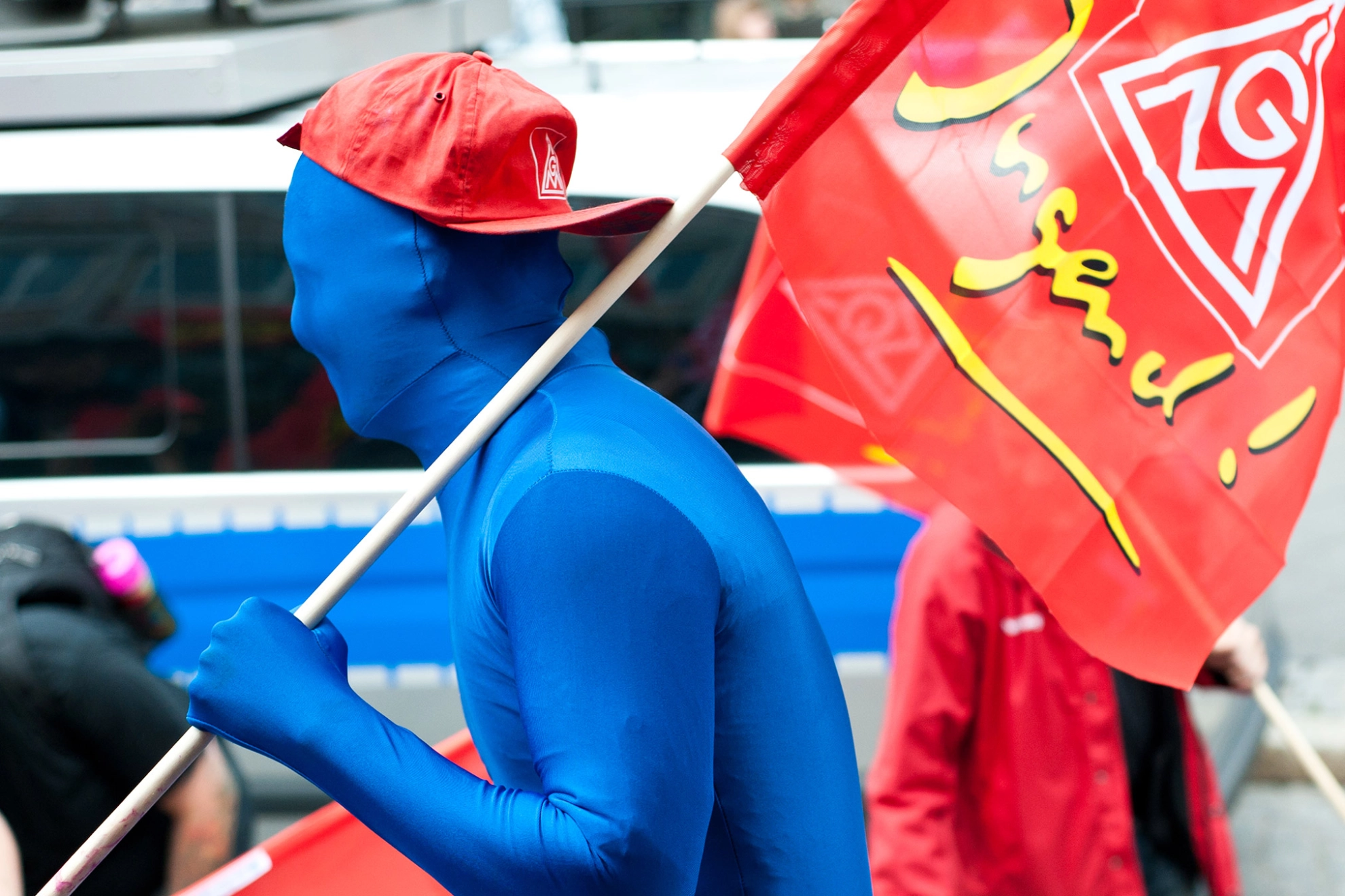 Junger Erwachsener mit blauem Morphsuit, IG Metall-Kappe und IG Metall Jugend-Fahne auf Demo.