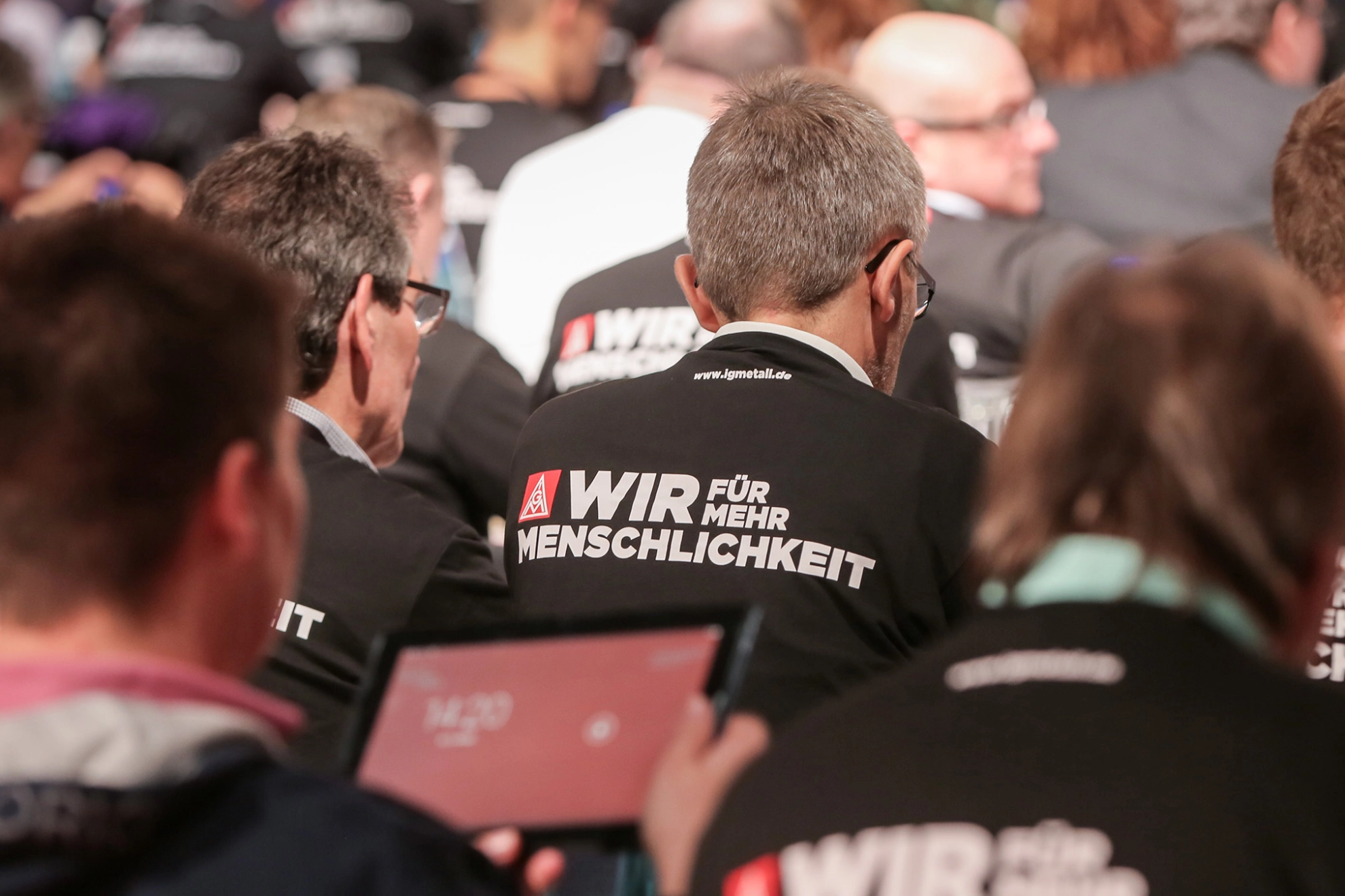 Sitzende Männer von hinten, auf deren Shirts "Wir für mehr Menschlickeit" steht.