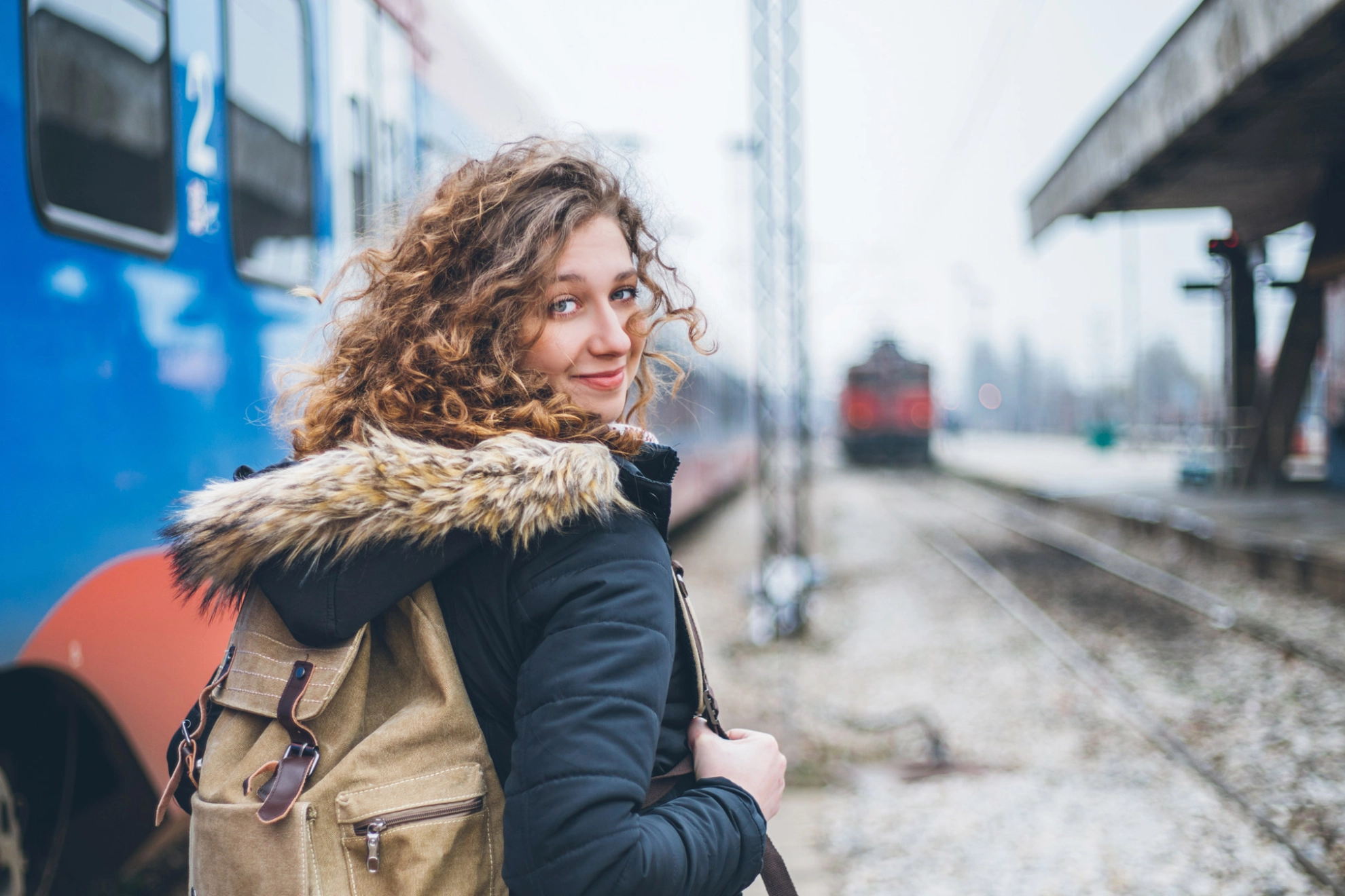 Junge Frau mit Rucksack auf dem Rücken steht vor einem Zug und blickt über ihre Schulter zurück in die Kamera.